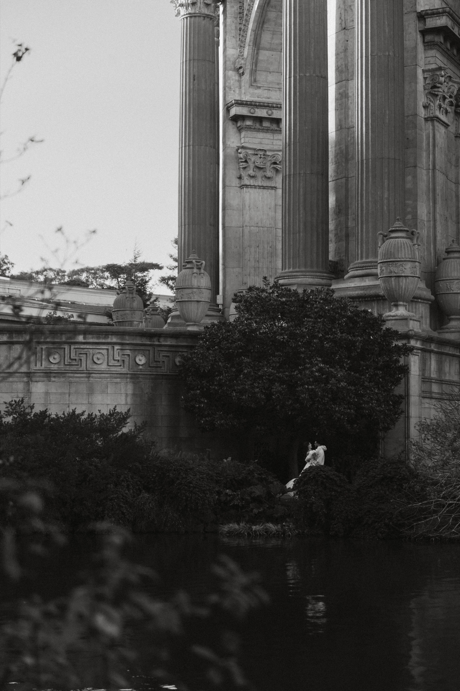 A distant black-and-white view of Berkeley and Robin beneath the Palace of Fine Arts colonnade, reflected in the lagoon